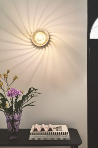 Lampa ścienna CHISELL Nordlux mosiądz zamontowana na ścianie nad stolikiem z kwiatami.