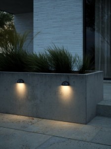 Lampa ścienna SCOR Nordlux 1xE14 15W Czarny zamontowana na betonowym murze oświetlającym zewnętrzną przestrzeń.