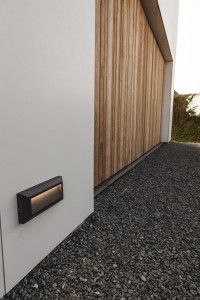 Kinkiet zewnętrzny LED Lutec Helena 5191602118 zamontowany na białej ścianie obok drewnianej elewacji domu.