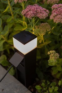 Słupek oświetleniowy Lutec Mains 7202201012 IP54 1xLED Aluminium Czarny w ogrodzie, w otoczeniu roślin.