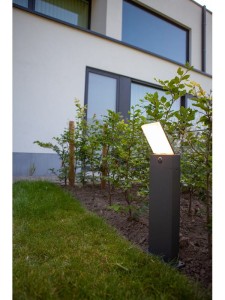 Słupek oświetleniowy Lutec Pano 7289001118 IP54 1xLED Aluminium na tle ogrodu przy nowoczesnym budynku.