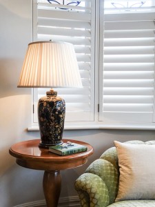 Lampa stojąca Black Birds Elstead Lighting na stoliku przy oknie z żaluzjami.